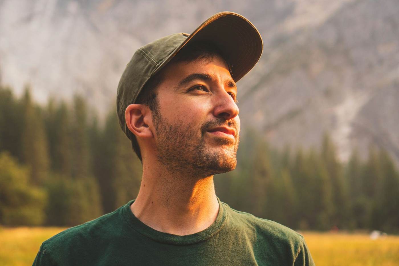 Man in a green cap and shirt looks up towards the sun with a happy expression.